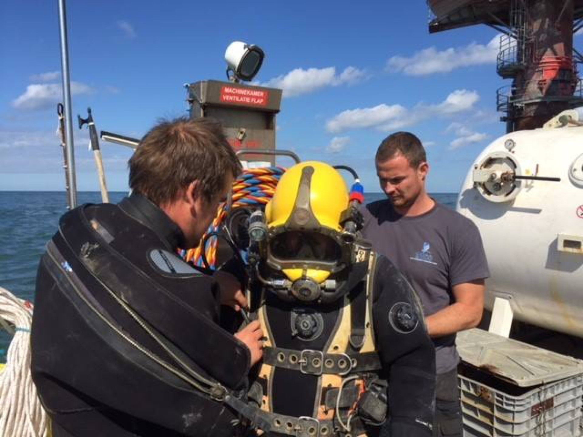 Two men assisting a diver in a yellow helmet and diving suit on a boat.