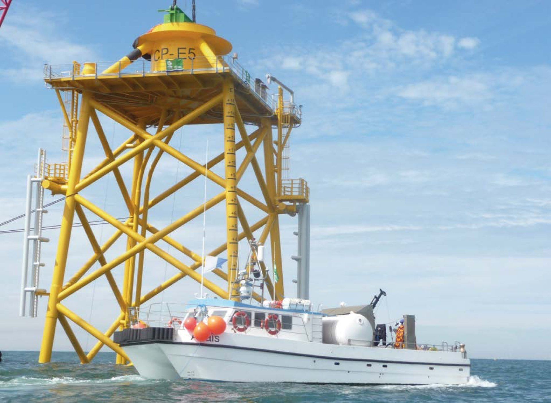 White boat near a large yellow offshore wind turbine platform in the ocean, blue sky.