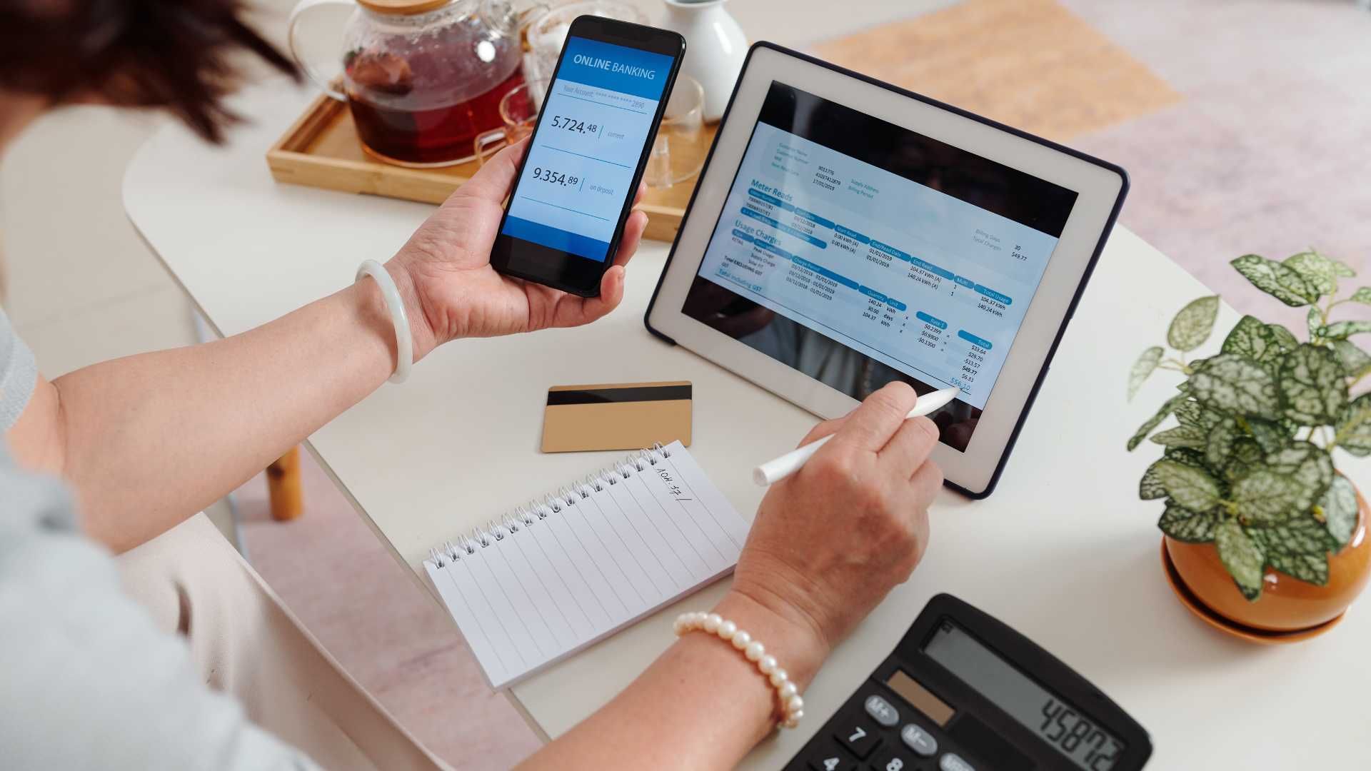 A woman is sitting at a table holding a cell phone and a tablet.