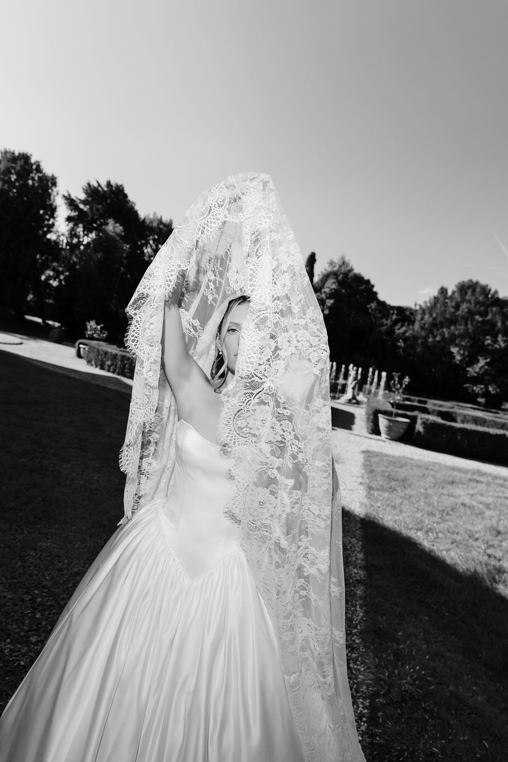 Black and white editorial portrait of a bride in a strapless satin ball gown lifting a scalloped lace veil over her head, peeking out with one eye, in the formal gardens of Villa Giona, during a luxury wedding in Italy