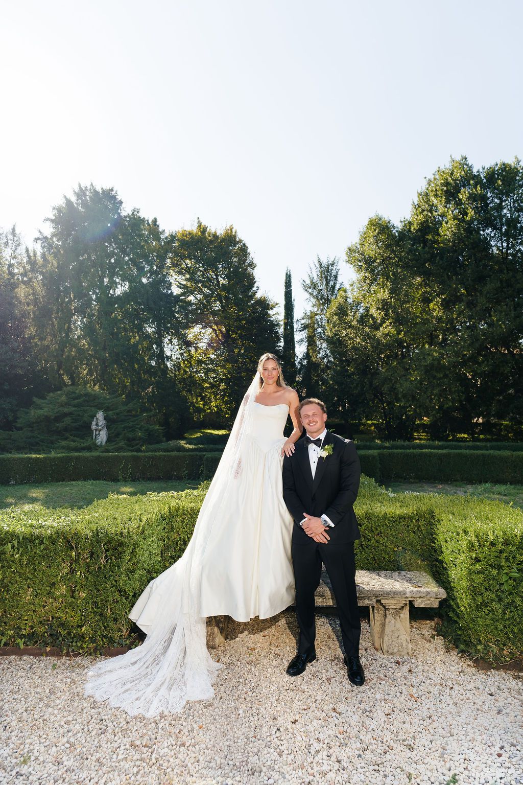 Bride in a strapless satin gown and long lace veil standing on a stone bench beside the groom in a tuxedo, in the formal boxwood garden of Villa Giona on a sunny day, during a luxury wedding in Italy