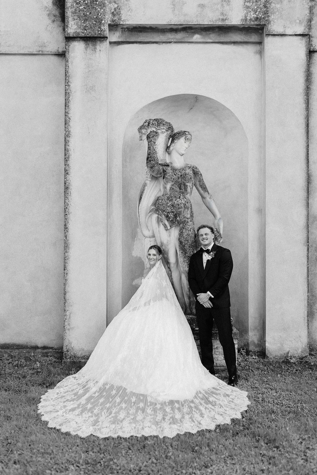 Black and white portrait of a bride with a long lace cathedral veil and a groom in a tuxedo standing before an antique stone statue in a niche at Villa Giona, during a luxury wedding in Italy