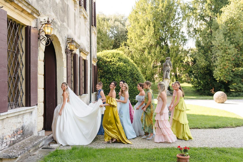 Bride in a strapless satin gown smiling over her shoulder at the entrance of Villa Giona while bridesmaids in mismatched colorful dresses carry her long veil, in the villa garden during a luxury wedding in Italy