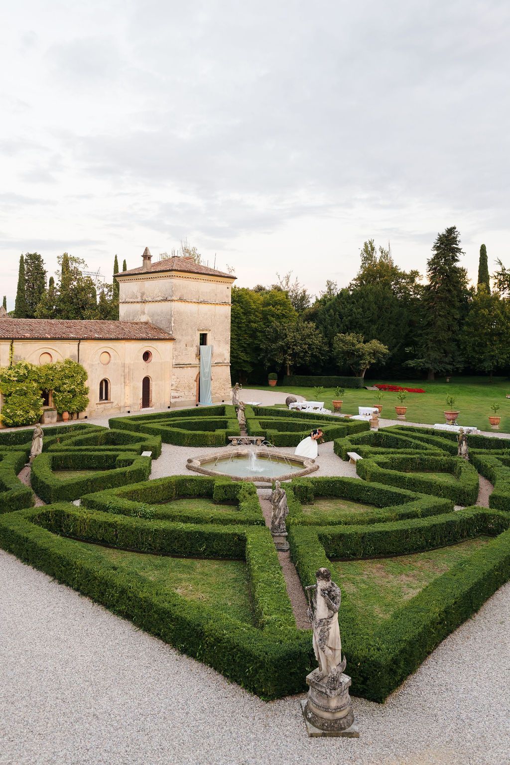Bride and groom kissing beside a fountain in the formal Italian garden of Villa Giona, with geometric boxwood hedges, antique stone statues and the historic villa tower, during a luxury wedding in Italy