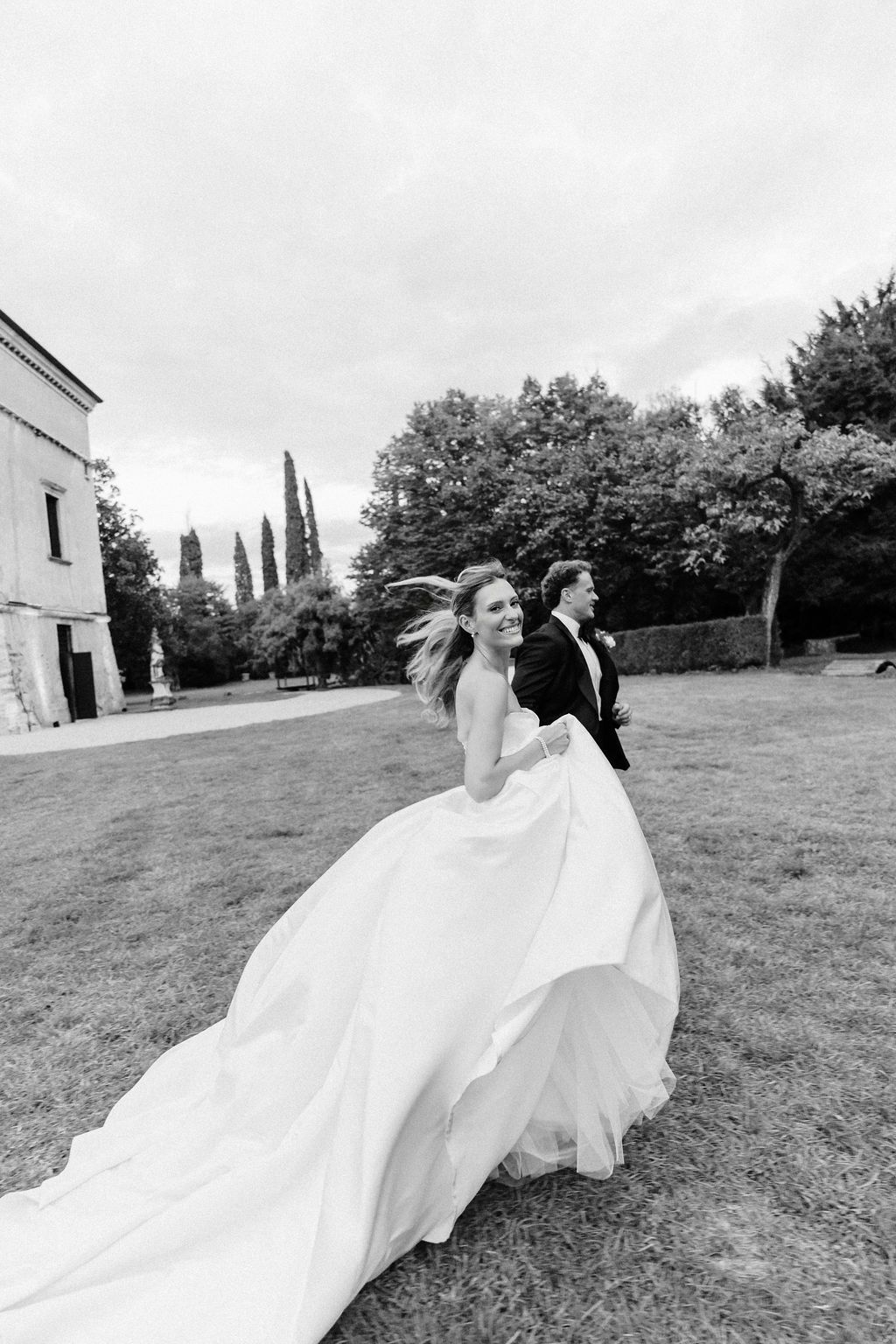 Black and white photo of a smiling bride running across the lawn with the groom in a tuxedo at Villa Giona, during a luxury wedding in Italy