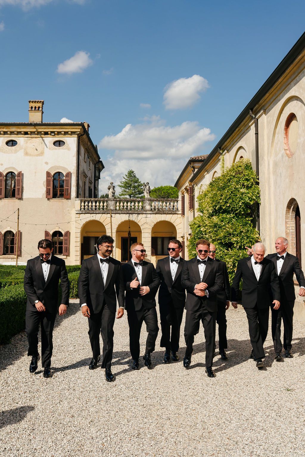 Groom and groomsmen in black tuxedos and sunglasses walking across the gravel courtyard of Villa Giona, with the historic villa, arched loggia and statues behind them, during a luxury wedding in Italy