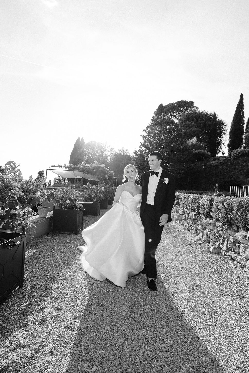 Black and white photo of a laughing bride in a strapless satin gown walking with the groom in a tuxedo along a backlit garden path with cypress trees at Villa Le Fontanelle, Florence, during a luxury wedding in Tuscany