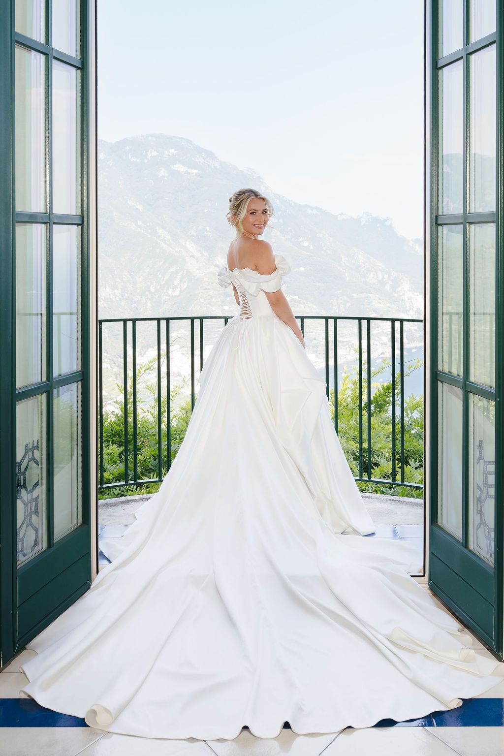 Bride in an off-the-shoulder satin ball gown with a long cathedral train standing on a balcony between green French doors, smiling over her shoulder with the Amalfi Coast mountains in the background, at Villa Eva in Ravello