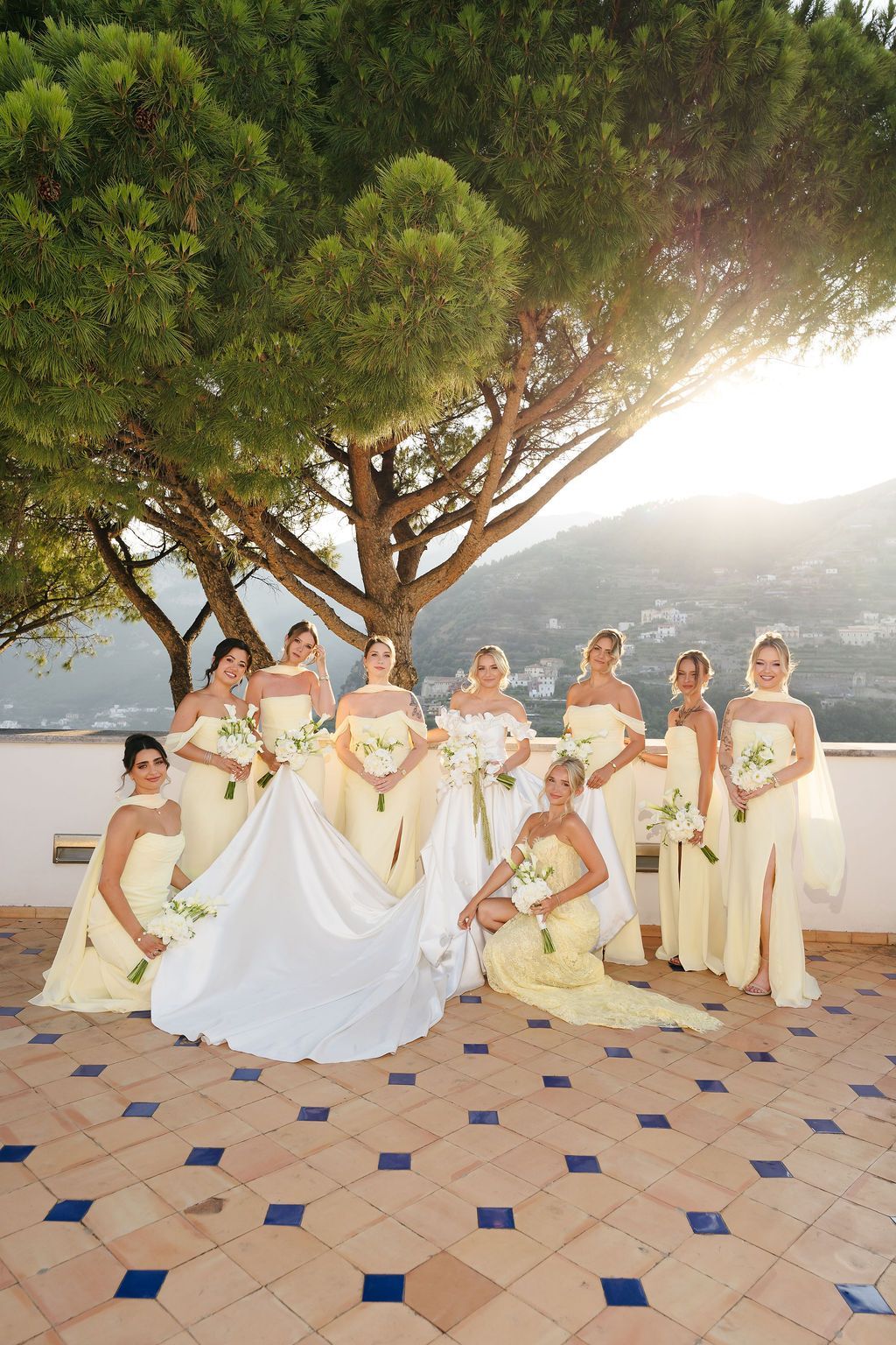 Bride in a white satin gown with her bridesmaids in pale yellow dresses holding white calla lily bouquets on a terracotta and blue tiled terrace under a stone pine at sunset, at Villa Eva in Ravello on the Amalfi Coast