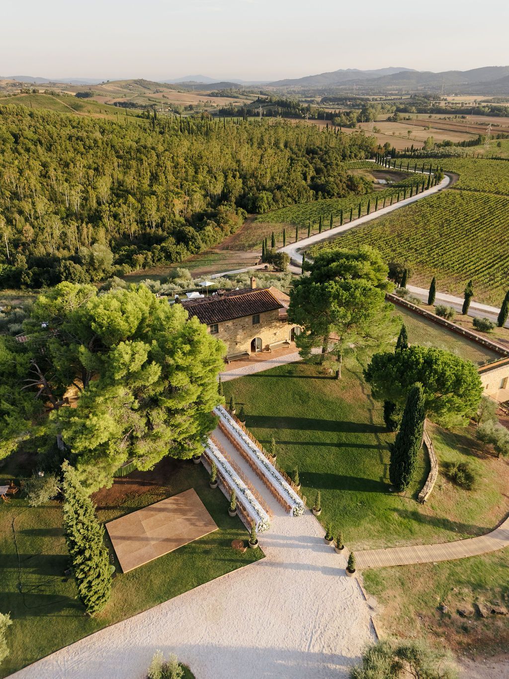Aerial drone view of an outdoor wedding dinner with long tables set beside a stone farmhouse, surrounded by vineyards, cypress-lined roads and rolling hills at Conti di San Bonifacio Wine Resort in Maremma, Tuscany