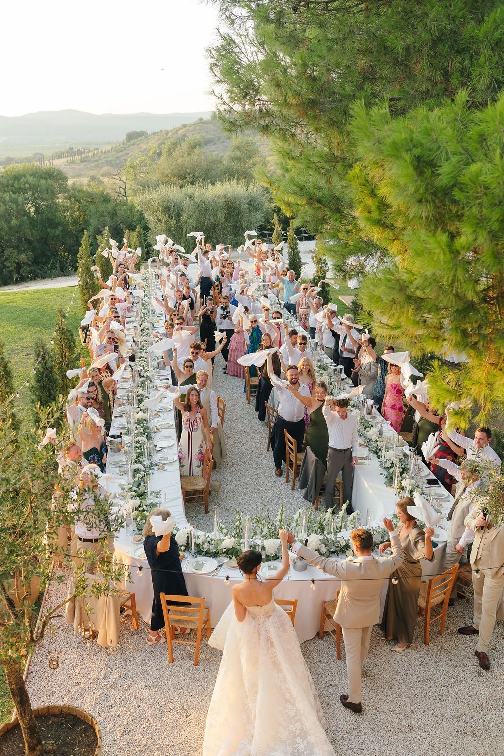 Bride and groom entering an outdoor wedding reception as guests wave white napkins around a long U-shaped table decorated with white flowers, at Conti di San Bonifacio Wine Resort in Maremma, Tuscany