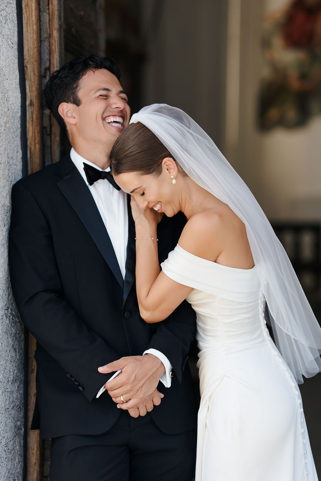 Candid photo of a laughing groom in a tuxedo and a smiling bride in an off-the-shoulder gown and veil leaning on his shoulder in a doorway at Villa Balbiano, Lake Como, during a luxury wedding in Italy