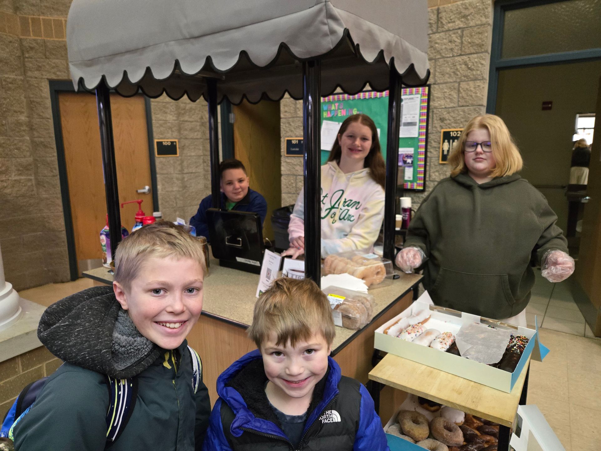 Children selling donuts from a stand in a hallway, smiling at the camera.