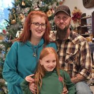 Family of three poses in front of a Christmas tree; woman in glasses, man with beard, and girl with red hair.