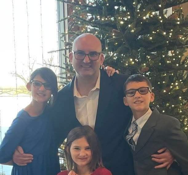 A man with three children pose near a decorated Christmas tree. They wear glasses and smile.