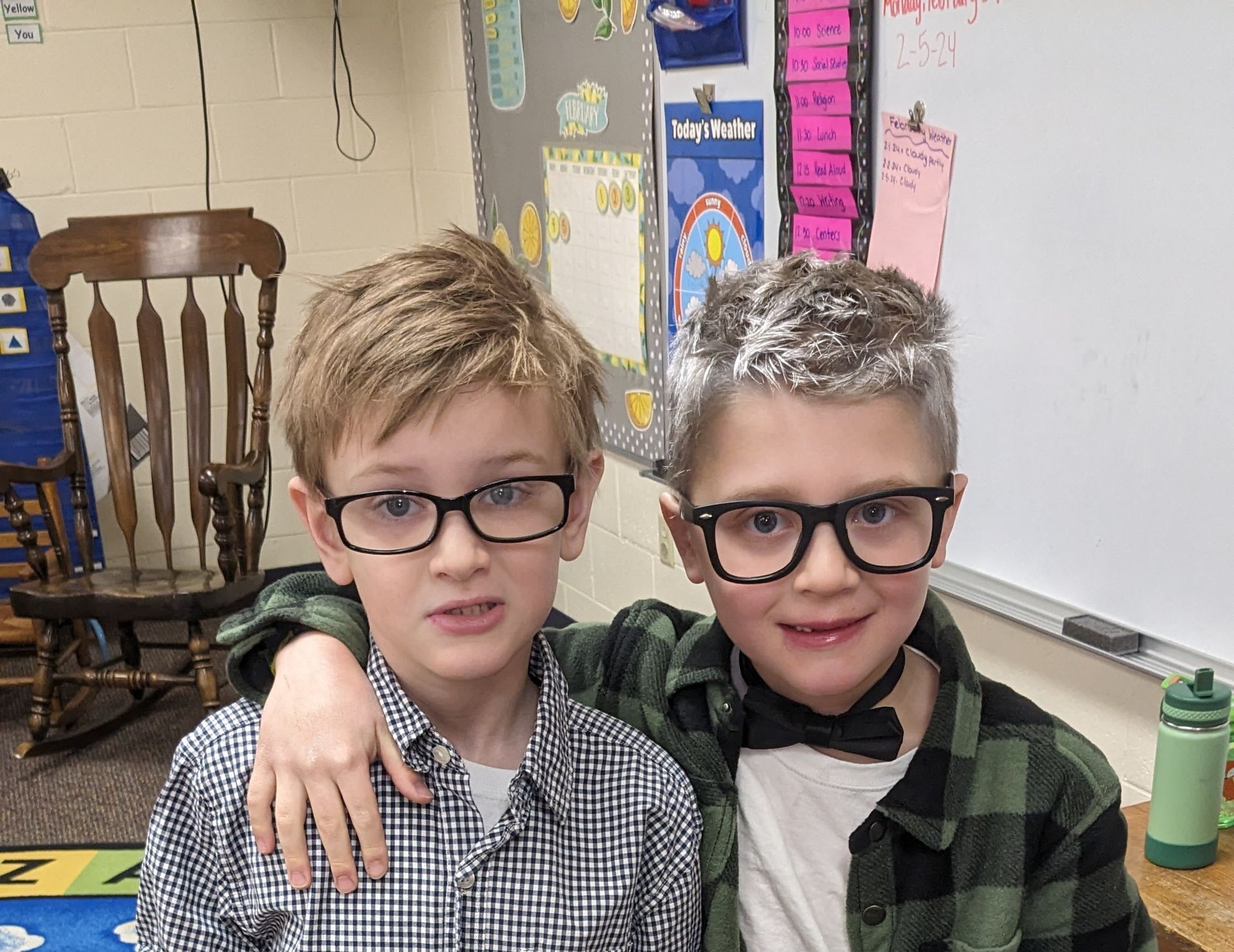 Two boys in glasses, one with white hair, pose in a classroom, one with an arm around the other.