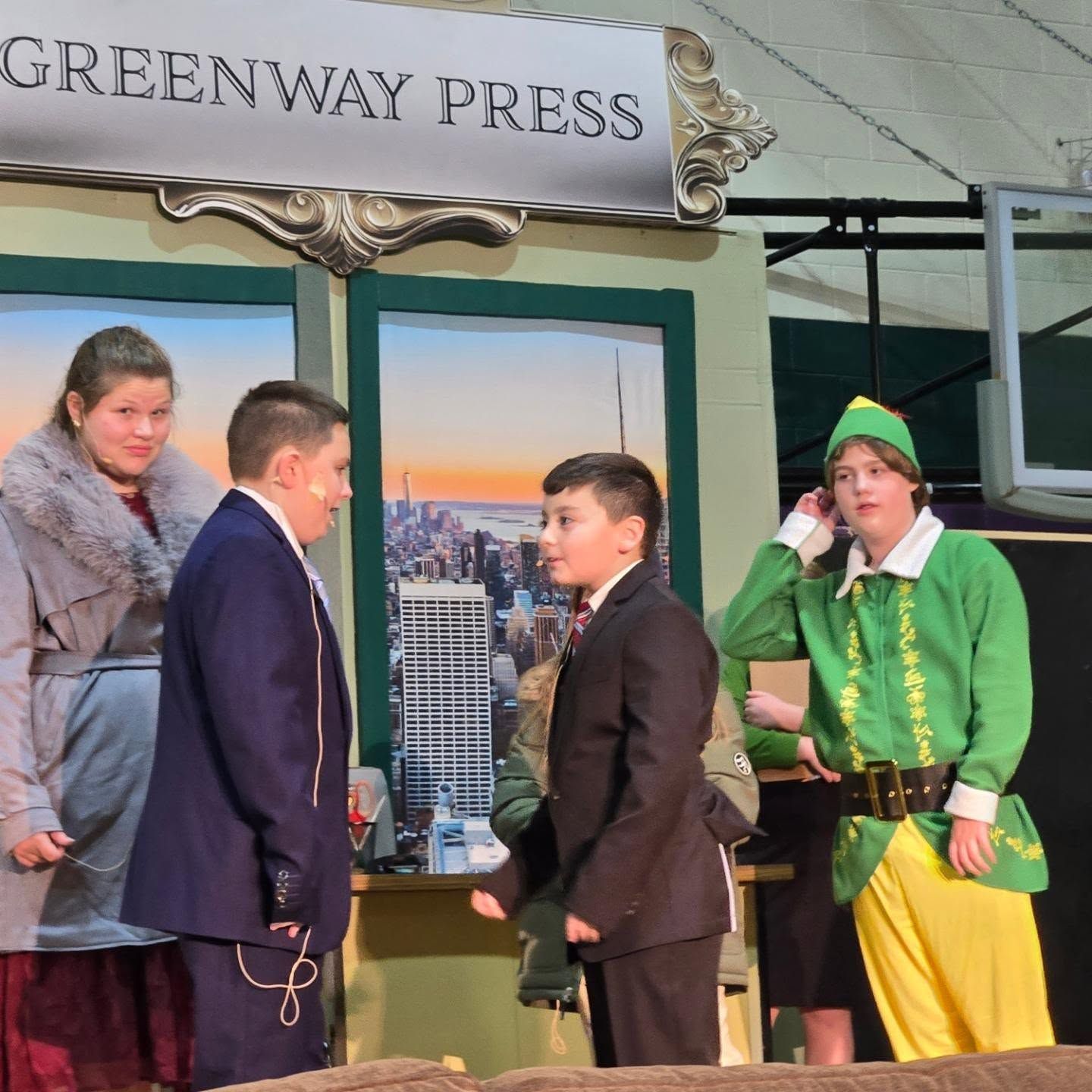 A stage play scene: Two boys in suits facing each other, actor in elf costume, woman in coat. 