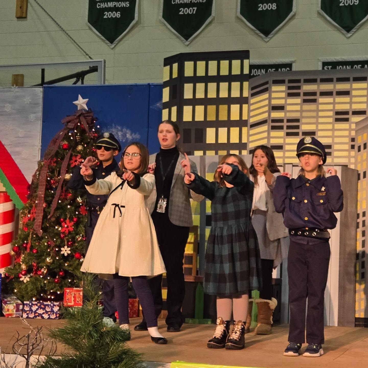 Children performing on stage with a Christmas tree, city backdrop, and one child dressed as a police officer.