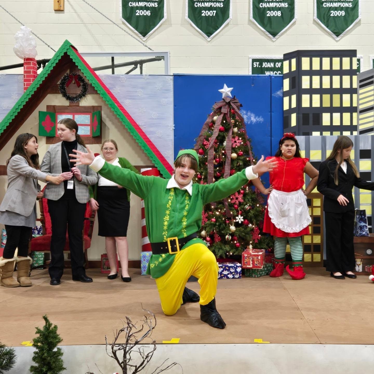 A stage production with a person dressed as an elf kneeling, arms outstretched, other characters in costume, and festive props.