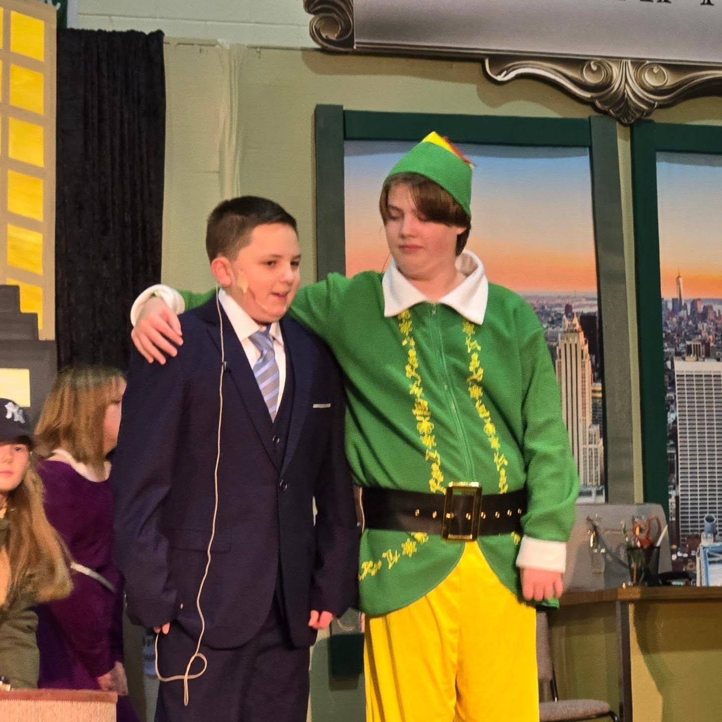 Two boys on stage; one in suit, one in elf costume, arms around each other. Stage background.
