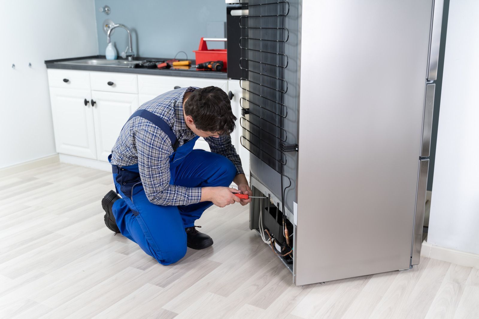 Technician crouches beside a dishwasher, repairing it in a bright kitchen.