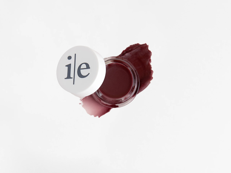 Open jar of burgundy-colored makeup with a swatch on white background.