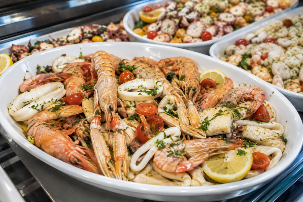 Una grande ciotola bianca con gamberetti e spicchi di limone su un vassoio da buffet, con altri piatti di pesce sullo sfondo.