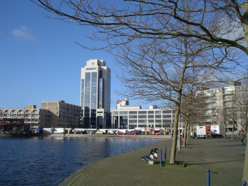 Een stel zit op een bankje bij een waterpartij met een hoog gebouw op de achtergrond