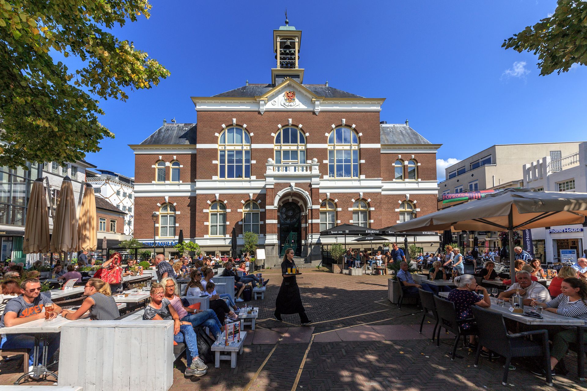 Een groep mensen zit aan tafels voor een groot bakstenen gebouw.