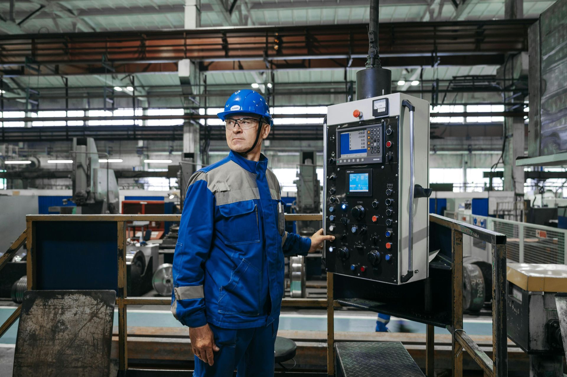 Un ouvrier en uniforme bleu et casque se tient à côté d'un panneau de commande industriel dans une usine.