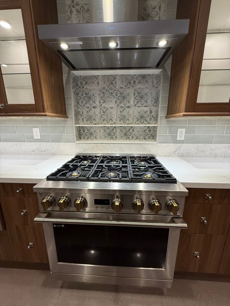 Stainless steel gas range with oven beneath a lit hood, centered in a modern kitchen with tile backsplash.