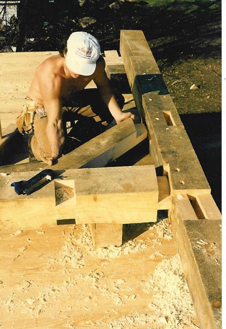 Man constructing timber frame. Wooden beams with mortise and tenon joints; sawdust on the floor.