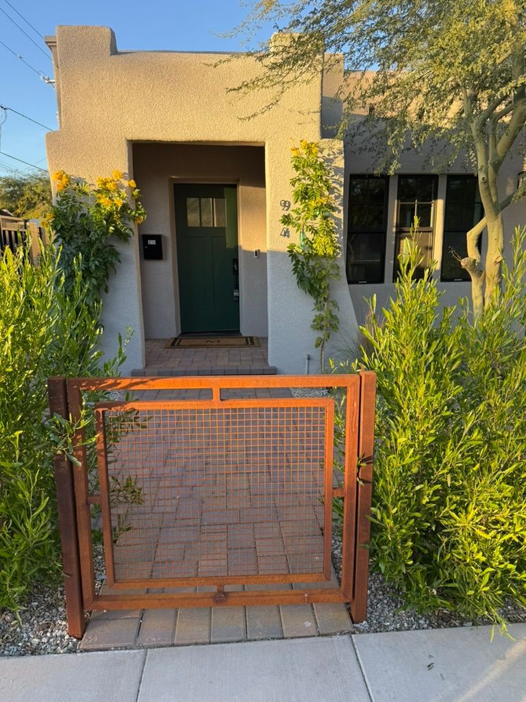 Small stucco house with a green front door, framed by dense shrubs and a rust-colored gate in front.