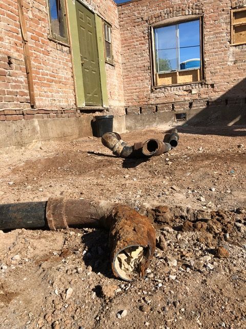 Pipes on dirt ground near a brick building. The lower pipe has a corroded end.