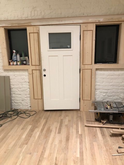 White door with glass pane, flanked by windows and wood trim, in a room with wood flooring.