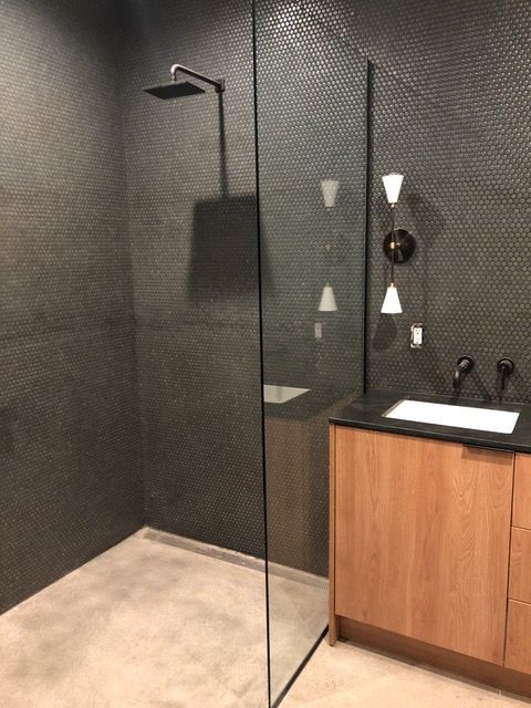Modern bathroom with dark gray tile walls, glass shower, wooden vanity, and concrete floor.