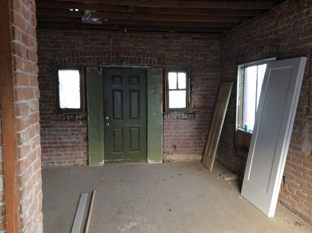 Interior view of a room with exposed brick walls, a green door, small windows, and construction materials.