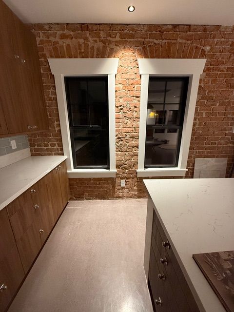 Kitchen with exposed brick wall, two windows with white trim, light cabinets, and a white countertop.