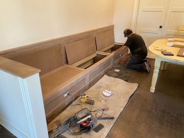 A person works on a built-in bench in a room. The bench is unfinished wood. Tools are scattered nearby.
