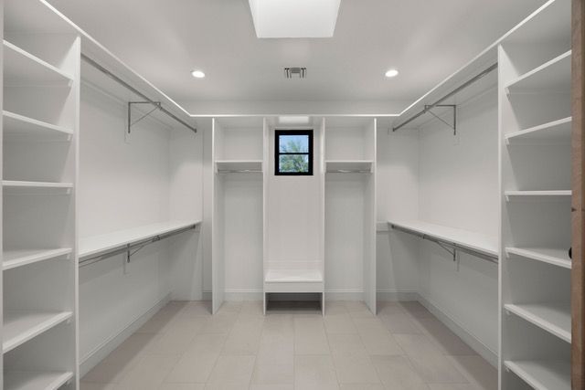 Empty, white walk-in closet with shelves and hanging rods. Has a small window at center, and a skylight.