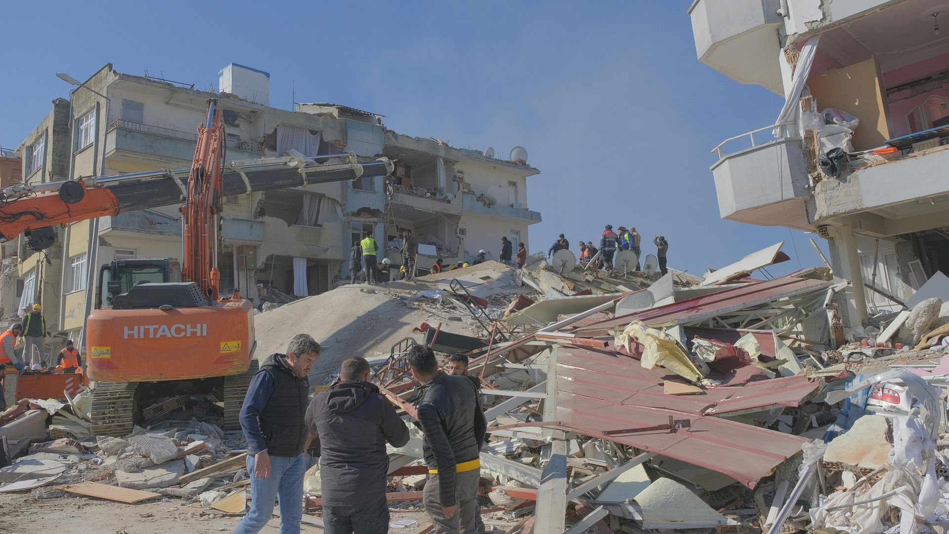 Collapsed buildings after an earthquake; rescue workers search debris under a bright blue sky.