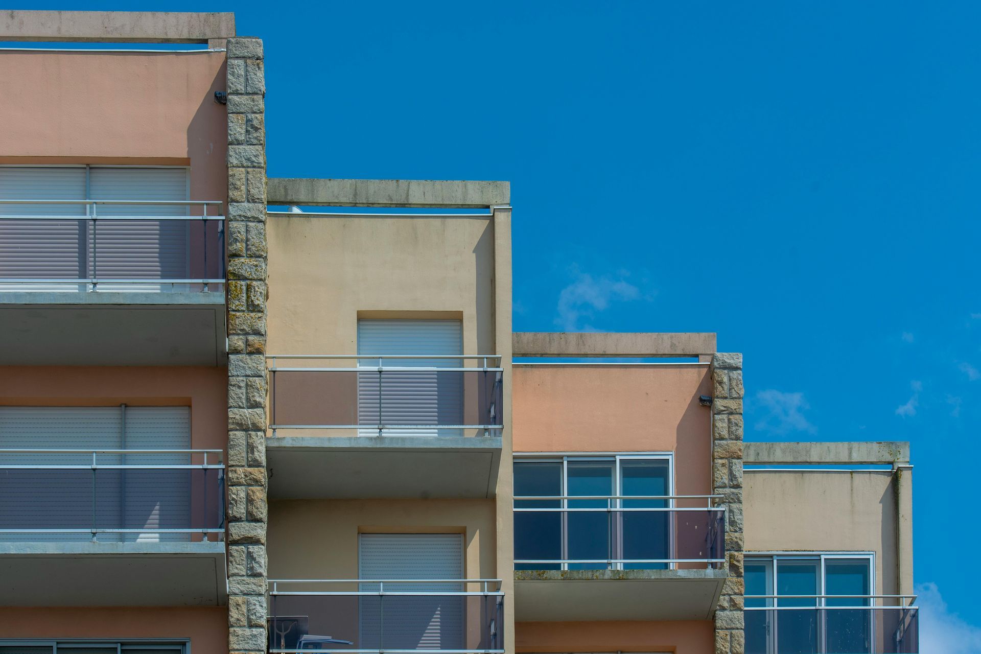 Apartment building with staggered balconies, tan and peach facade, blue sky backdrop.