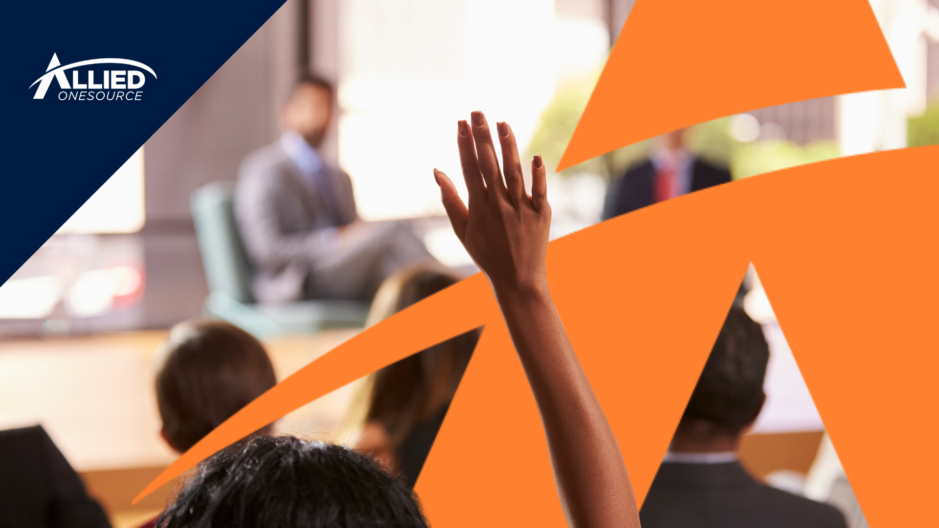 A person is raising their hand in a conference room setting with attendees listening to a speaker se