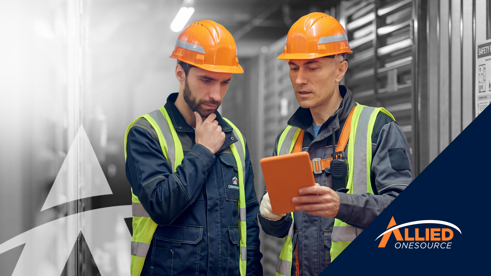 Two industrial workers in orange hard hats and safety vests review a tablet together. 