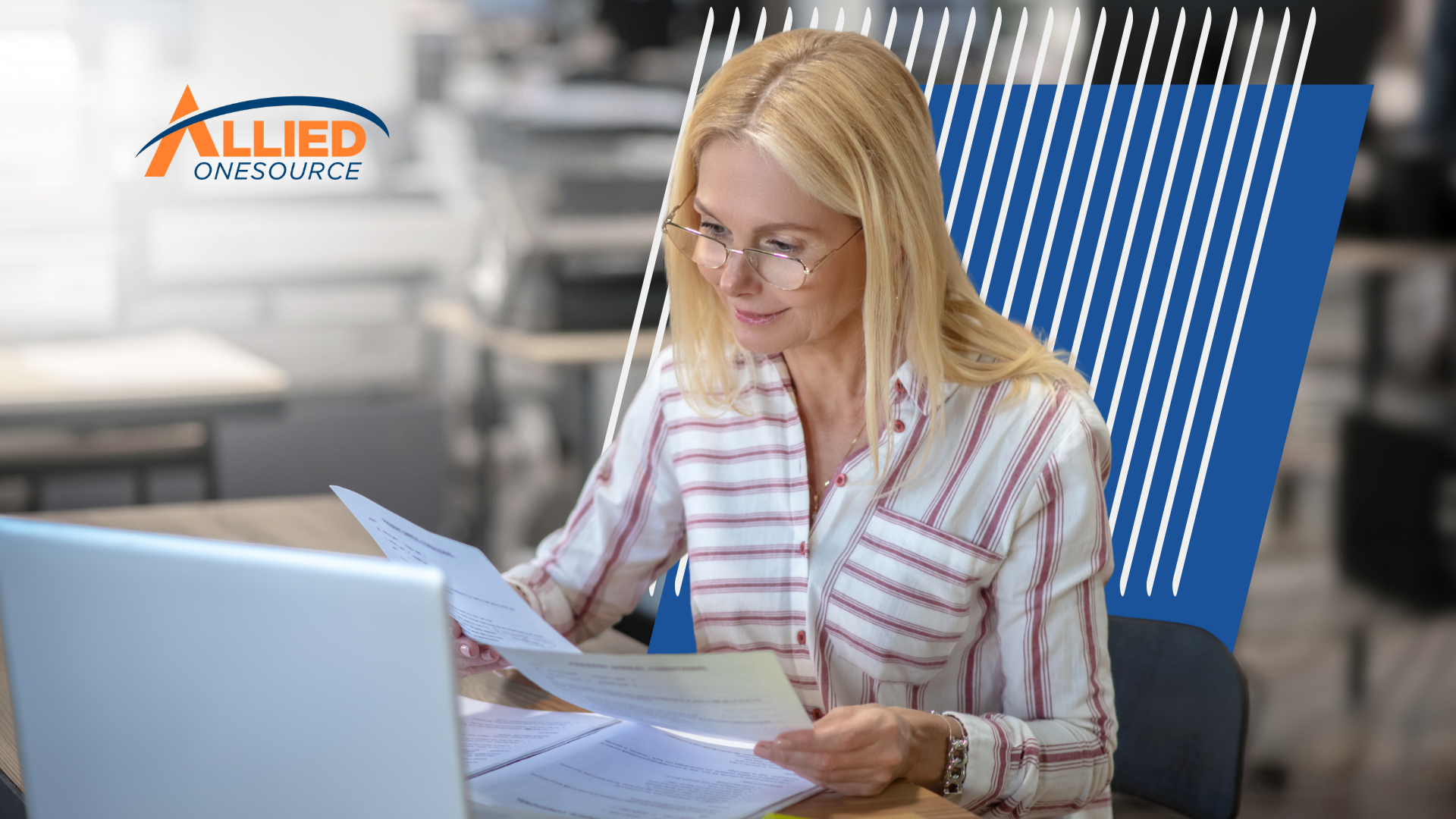 A woman in glasses reviews printed documents at her desk next to an open laptop.