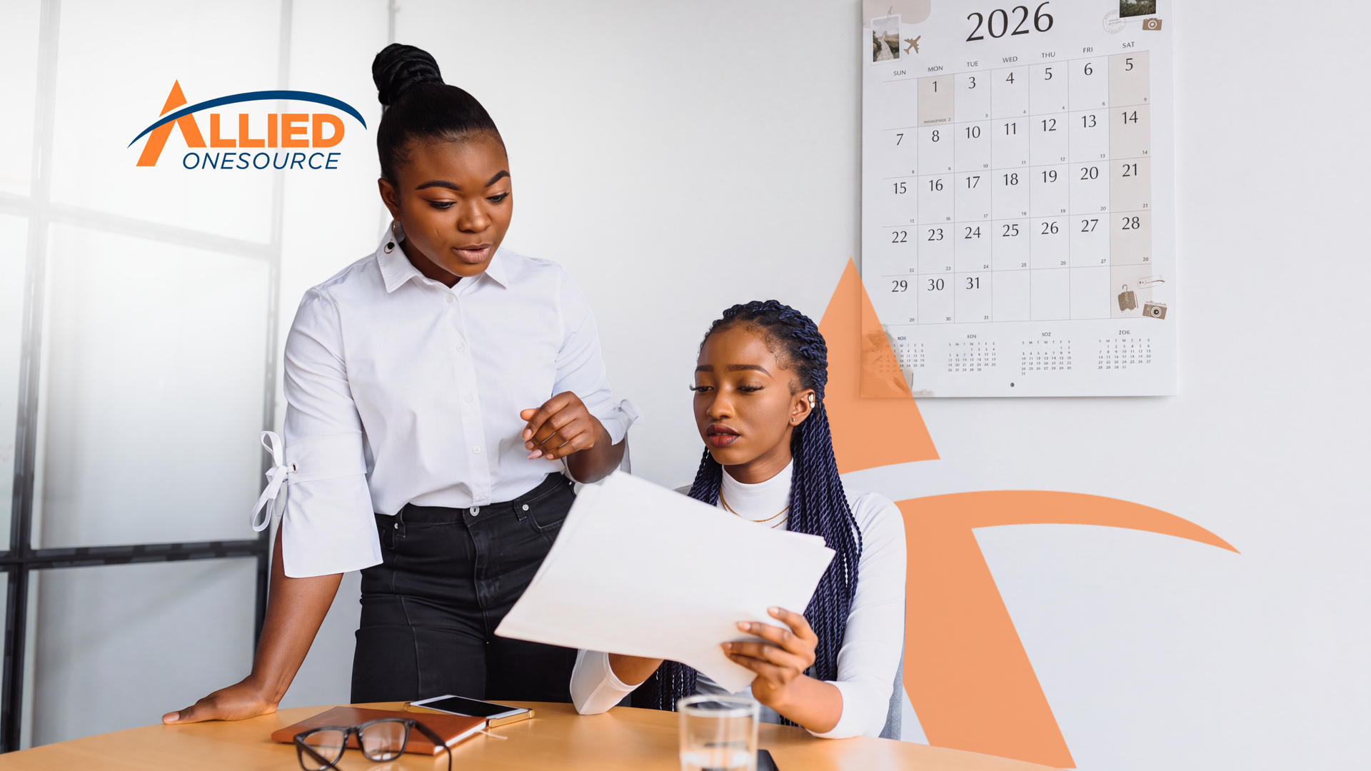 Two women review printed documents together at a table in a bright office, with a 2026 wall calendar