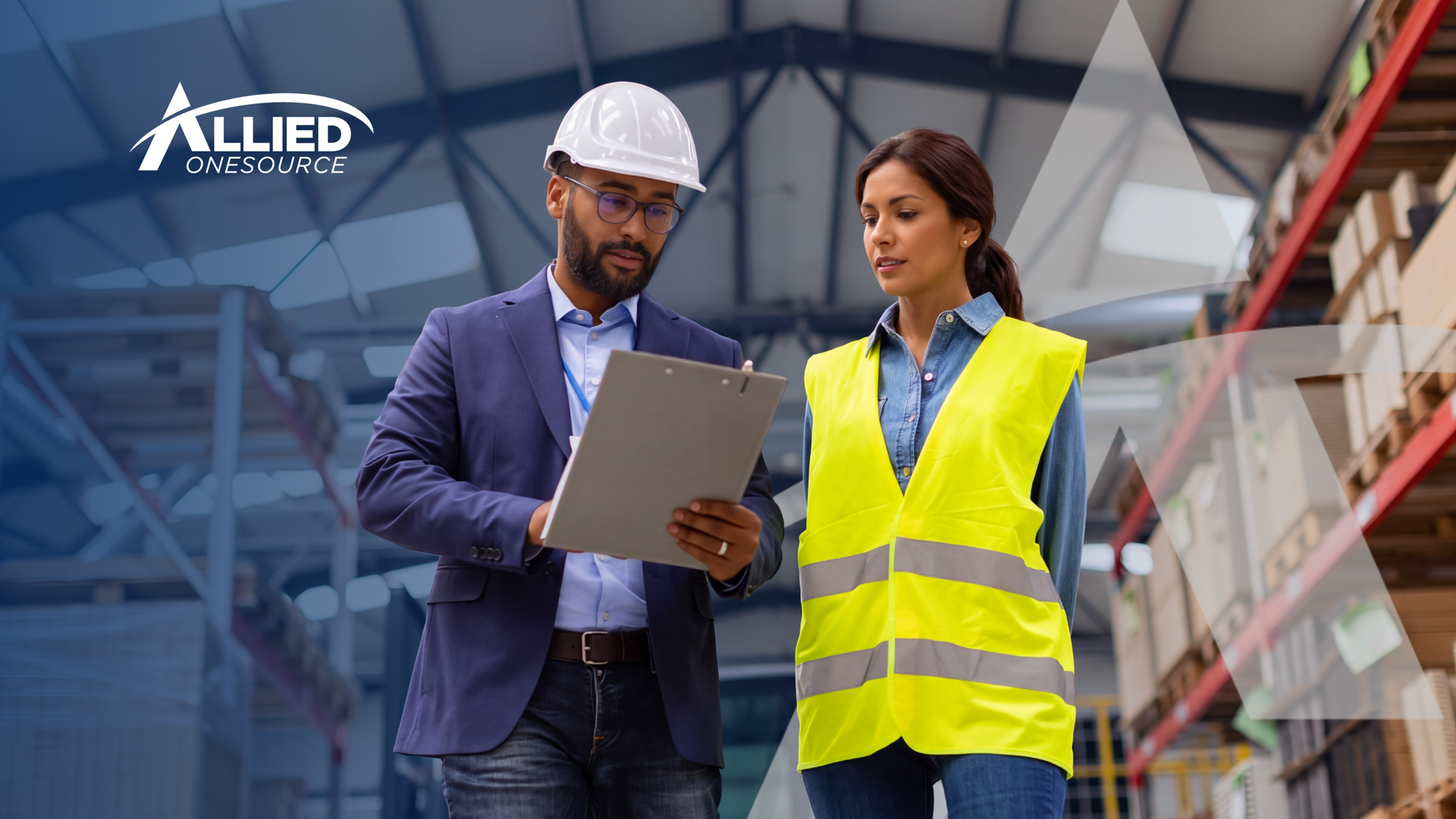 A man in a hard hat and a woman in a safety vest walk through a warehouse reviewing a clipboard. All