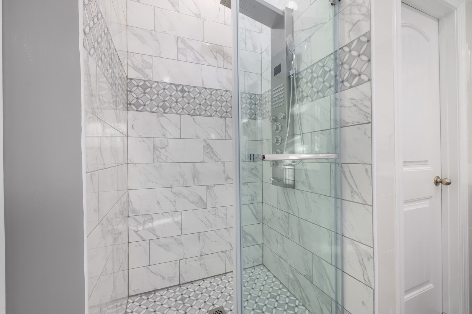 Modern white tiled shower with glass enclosure and small shelving, beside a white door