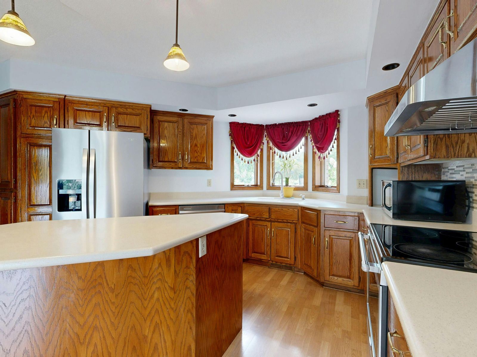Bright kitchen with wood cabinets, stainless appliances, white counters, and red curtains by the window