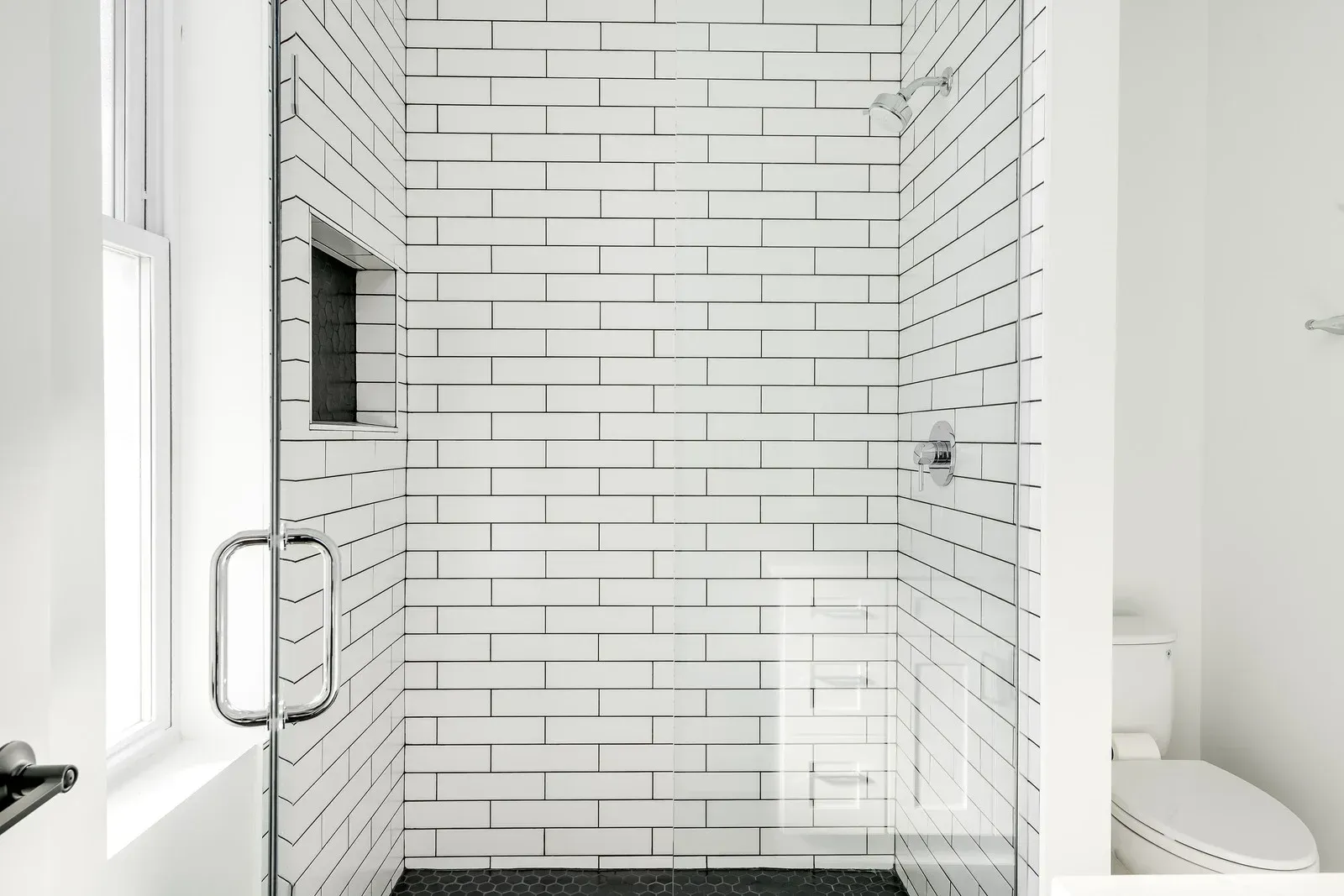 Modern white-tiled walk-in shower with glass door and black floor tile in a bright bathroom
